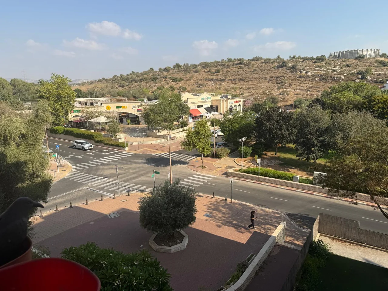צילום מהחלון של צומת רחוב עם חנויות, עצים, מכוניות וגבעה סלעית ברקע
